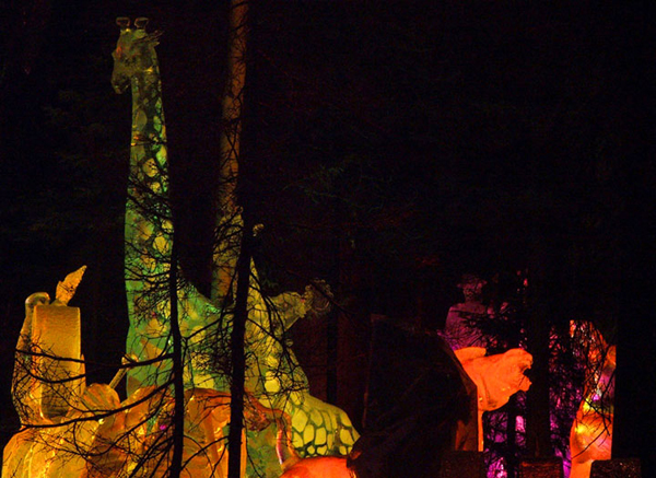 Animal Parade ice sculpture, lit by colored lights at night. Seen through the trees. By Steve Brice, Heather Brown, Tjana Raukar, and Mario Amegee. Ice Alaska 2005